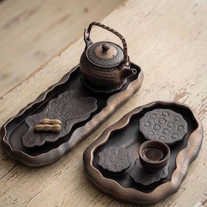 May include: A dark brown ceramic tea set featuring a teapot, two trays, and a cup. The teapot has a woven handle and rests on a tray with a leaf design. The second tray has a lotus seed pod design and a small cup. The set includes a pair of peanuts.