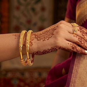 May include: Close-up of a hand adorned with gold jewelry, including two ornate bangles and a ring. The hand is decorated with henna designs. The sleeve of a magenta garment with gold trim is visible in the background. The jewelry is likely made of gold.