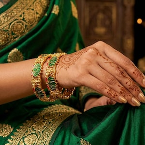 May include: Gold bangles with green and red accents adorn a wrist, set against a backdrop of a green sari with gold embroidery. The intricate design of the bangles complements the henna-decorated hand, showcasing traditional Indian jewelry.