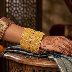 May include: Two ornate gold bangles with intricate floral patterns adorn a wrist. The arm is clad in a maroon silk sleeve with gold trim. The hand is decorated with henna. The bangles rest on a blue and gold patterned fabric.