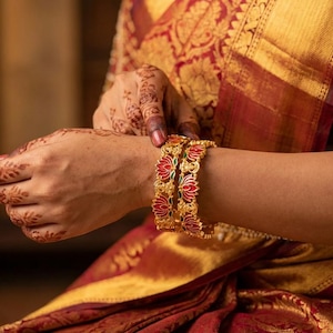 May include: Close-up of gold bangles with red and green floral designs. The bangles are worn on a wrist, with intricate henna patterns on the hand. The person is wearing a red and gold sari with embroidered sleeves.