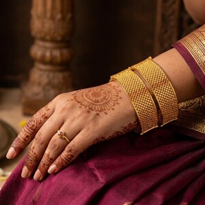 Op de afbeelding: Close-up van een hand versierd met henna en gouden sieraden. Twee getextureerde gouden armbanden omcirkelen de pols, aangevuld met een delicate ring. De hand rust op een diep bordeauxrode stof, met een goudgeborduurde rand zichtbaar.