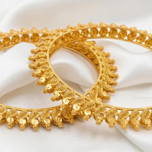 May include: Two gold bangles with intricate leaf-like designs. The bangles are displayed on a white, silky fabric. The jewelry has a bright, polished finish, reflecting light. The overall theme is elegant and ornate.
