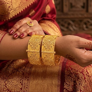 May include: A person's arm displays two elaborate gold bangles, each with detailed designs. The bangles are set against a red and gold patterned fabric. A gold ring with a red gemstone is also visible. The image is rich in detail.