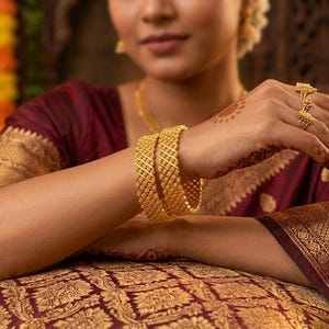 Op de afbeelding: Close-up van gouden sieraden, waaronder een ketting, ringen en twee dikke armbanden. De armbanden hebben een traliemotief. De persoon draagt een bordeauxrode en goudkleurige sari. De achtergrond is wazig, met warme verlichting.