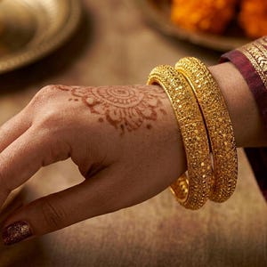Könnte beinhalten: Nahaufnahme von zwei goldenen Armreifen mit aufwendigen Mustern. Die Hand ist mit Henna-Kunst verziert und in einen bordeauxroten und goldenen Stoff gehüllt. Ringelblumen und eine goldene Platte sind im Hintergrund.