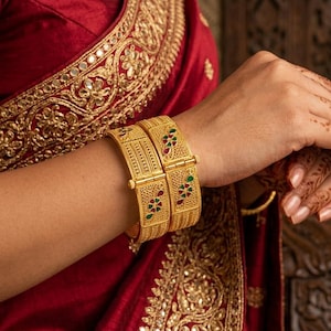 Pode incluir: Close-up de pulseiras douradas com designs intrincados, adornadas com detalhes vermelhos e verdes. As pulseiras são usadas no braço de uma pessoa, complementadas por uma peça de roupa vermelha com bordados dourados. A imagem mostra joias tradicionais.
