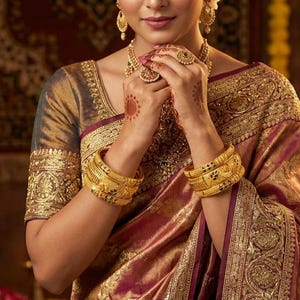 May include: A woman wearing a traditional Indian sari in shades of gold and burgundy. The sari is richly embroidered with gold thread. She is adorned with gold jewellery, including earrings, a necklace, rings, and bangles.