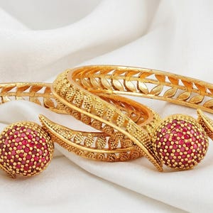 May include: Gold-coloured bangles and matching earrings with a red gemstone cluster design. The bangles feature intricate leaf-like cutouts and detailed patterns. The jewellery is displayed on a white, textured fabric.