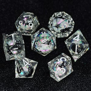 Könnte beinhalten: Ein Set aus sieben polyedrischen Würfeln aus klarem Harz, die jeweils irisierende Glitzerpartikel und kleine schwarze Katzenillustrationen enthalten. Die Zahlen sind in einer kräftigen, silberfarbenen Schrift auf den Außenflächen aufgedruckt.