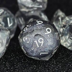 Könnte beinhalten: Ein zwanzigseitiger Würfel mit einer facettierten, transparenten Außenhülle und einem dunklen, glitzernden Kern. Der Würfel zeigt eingravierte weiße Zahlen, darunter eine deutliche neunzehn, die auf der geometrischen Oberfläche verteilt sind.