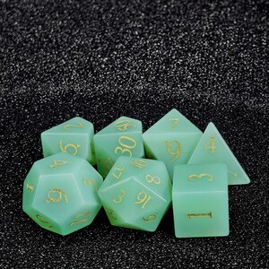 Könnte beinhalten: Ein Satz von sieben Würfeln in verschiedenen geometrischen Formen, darunter ein d4, d6, d8, d10, d12, d20 und ein Prozentwürfel. Die Würfel sind transluzent seegrün mit goldfarbenen Zahlen. Die Würfel sind für Tabletop-Spiele.
