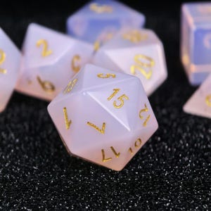 Op de afbeelding: Close-up van verschillende polyhedrale dobbelstenen. De dobbelstenen zijn doorschijnend wit met een roze tint en gouden cijfers. De dobbelstenen zijn voor tabletopspellen zoals Dungeons and Dragons.