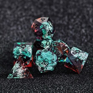 Könnte beinhalten: Ein Set aus polyedrischen Würfeln aus durchscheinendem Kunstharz mit eingebetteten smaragdgrünen und tiefroten Pigmenten, akzentuiert durch schimmernde Silberfolienflocken. Jeder Würfel zeigt klare, schwarz eingefärbte Ziffern.