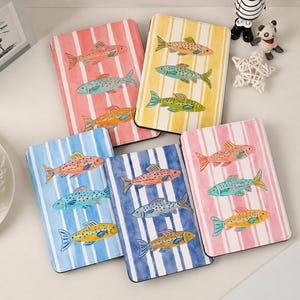 Puede incluir: Cuadernos con ilustraciones de peces coloridos sobre fondos a rayas. Los cuadernos presentan diseños de peces en tonos naranja, azul y amarillo. Los fondos están a rayas en rosa, amarillo, azul y rojo.