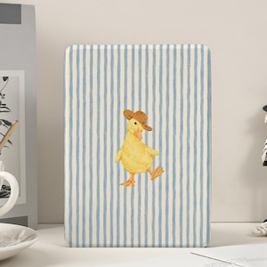 Op de afbeelding: Een rechthoekige hoes met een blauw en wit gestreept patroon. De hoes heeft een aquarel illustratie van een gele eend met een bruine cowboyhoed. De eend loopt met gespreide benen. De hoes staat op een wit oppervlak.