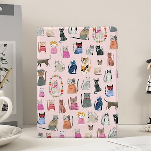 May include: A pink tablet case featuring a repeating pattern of various cartoon cats. The cats are in different poses and colors, some wearing clothes. The case has a rounded top and is set against a white background.