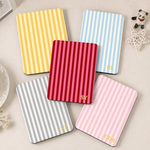 Puede incluir: Cinco cuadernos rectangulares con cubiertas de rayas verticales en amarillo, azul claro, rojo oscuro, gris y rosa claro. Los cuadernos rojo, azul, gris y rosa tienen iniciales de monograma doradas impresas en la esquina inferior derecha.