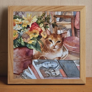 May include: Watercolor painting of a ginger cat resting near a vase of flowers and a stack of books. The artwork is framed in a light wood frame. The flowers include yellow sunflowers and red roses. The cat is looking directly at the viewer.
