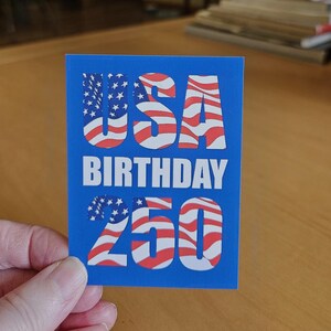 May include: A rectangular sticker with a solid blue background featuring the text USA in a bold font filled with a red, white, and blue American flag pattern, the word BIRTHDAY in white, and the number 250 with the same flag pattern fill.