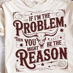 Może przedstawiać: Kremowy t-shirt z krótkim rękawem z ciemnobrązowym napisem IF I'M THE PROBLEM, THEN YOU MIGHT BE THE REASON. Wzór zawiera centralną ilustrację czaszki byka, ozdobne zawijasy oraz małe gwiazdki rozmieszczone wokół głównego motywu graficznego.