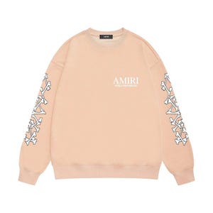May include: A pale peach sweatshirt featuring white bone patterns on the sleeves. The text "AMIRI HOLLYWOOD.CA" is printed on the chest. The sweatshirt has a crew neck and long sleeves.