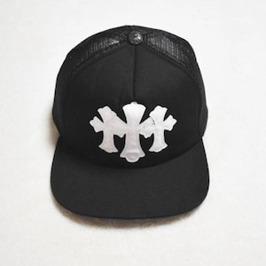 Puede incluir: Gorra de camionero negra con un diseño de cruz plateada en la parte delantera. La gorra tiene una parte trasera de malla y una visera curva. Un pequeño botón plateado está en la parte superior.
