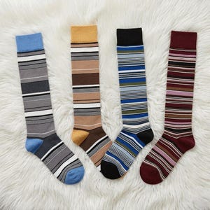May include: Four pairs of knee-high socks with horizontal stripes in various colour combinations. The socks include blue, grey, brown, and burgundy with stripes of white, black, and other colours. The socks are displayed on a white, fluffy surface.