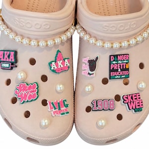 May include: A pair of light pink clogs adorned with white pearl straps and various colorful charms. The charms feature text including AKA, 1908, Skee Wee, and Living Pretty, along with a hand gesture icon and a sign reading Danger Pretty & Educated.