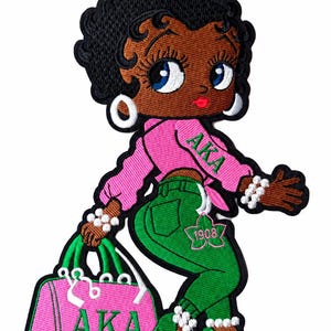 May include: An embroidered patch featuring a cartoon character with black hair, wearing a pink long-sleeve top with the green text AKA, green pants with a pink ivy leaf and 1908, and green high heels. The character holds a pink bag with AKA girl.