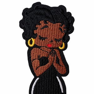 May include: An embroidered patch featuring a stylized character with dark skin, curly black hair, and gold hoop earrings. The character wears a black strapless dress with white side accents, has red lips, and holds hands together near the chest.