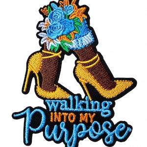 May include: An embroidered patch featuring dark brown legs wearing yellow high-heeled shoes with ankle straps. A bouquet of blue and orange flowers rests above the ankles. The text "walking INTO MY Purpose" is embroidered in blue and orange below.
