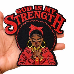 May include: An embroidered patch featuring a person with an afro hairstyle wearing a red headband and red hoodie, hands clasped in prayer, with gold hoop earrings and a cross necklace. The text GOD IS MY STRENGTH is arched in red above the figure.