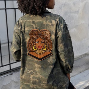 May include: A camouflage print jacket featuring a large embroidered patch on the back. The patch depicts a person with brown skin, textured hair styled in two buns, gold hoop earrings, and a gold necklace, set against a dark brown background.