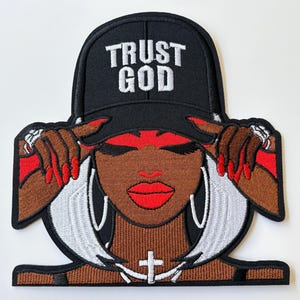 May include: An embroidered patch featuring a person with brown skin, red lips, and white hair wearing a black baseball cap with the white text TRUST GOD. The person has hands with red nails near the cap, silver hoop earrings, and a cross necklace.