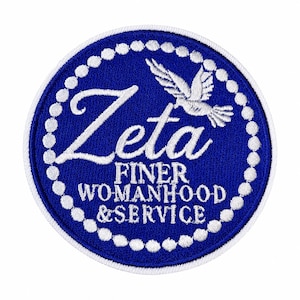 May include: A circular embroidered patch with a royal blue background and a white border of small circles. The center features white text reading Zeta FINER WOMANHOOD & SERVICE, accompanied by a white dove graphic positioned above the text.