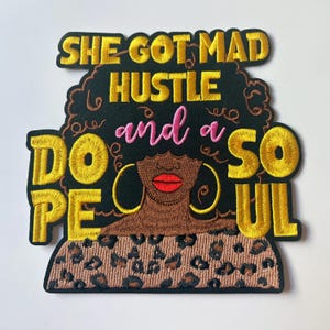 May include: An embroidered patch featuring a stylized illustration of a person with dark skin, red lips, and large gold hoop earrings. The text reads SHE GOT MAD HUSTLE and a DOPE SOUL in yellow and pink thread against a black background.