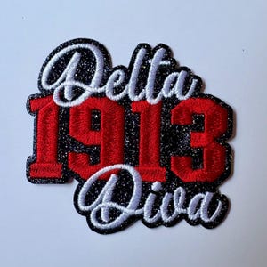 May include: A black glittery embroidered patch featuring the white script text Delta and Diva with the red block numbers 1913 centered between the words. The design has a thick black border and a textured surface with a sparkling finish.