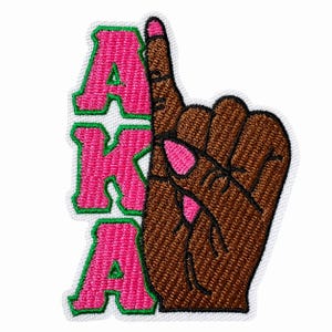 May include: An embroidered patch featuring the letters AKA in bright pink with a green border, positioned next to a brown hand with a raised index finger and pink fingernails. The design is outlined in white thread on a white fabric backing.