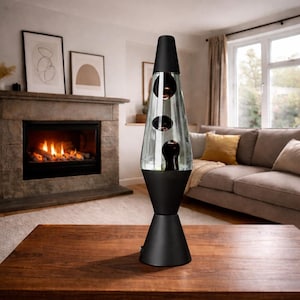 May include: A black lava lamp with a clear glass body containing dark red lava. The lamp has a conical base and top, and is placed on a wooden table. The background includes a fireplace and a living room setting.
