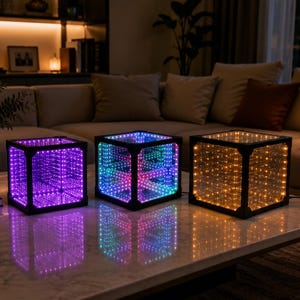 May include: Three illuminated infinity cube lamps with black frames on a marble table. The lamps display purple, multicoloured, and amber lights. These lamps are a modern home decor item.