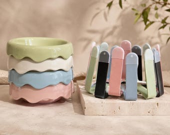 May include: A stack of four ceramic bowls with wavy, scalloped rims in pastel green, white, light blue, and pink. Beside the stack is a circular metal rack featuring upright, U-shaped dividers in matching pastel colors and black, arranged in a ring.