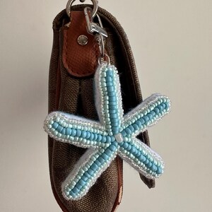 Puede incluir: Un bolso de mano marrón con monograma, correa de cadena plateada y un dije en forma de estrella de mar. El dije está elaborado con cuentas azul claro y blancas, tiene un respaldo de fieltro blanco y se sujeta con un mosquetón de metal plateado.