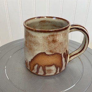 May include: A ceramic mug featuring a brown horse silhouette. The mug has a speckled beige and brown exterior with a brown handle. The interior rim and handle stripes are also brown. The mug is on a grey surface.
