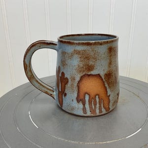 May include: A handmade ceramic mug with a light blue glaze and brown accents. The mug features a brown illustration of a desert animal and cactus. The mug has a large handle and is suitable for hot or cold beverages.