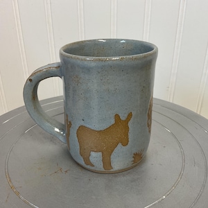 Peut inclure: Une tasse en céramique faite à la main avec un glaçage bleu clair. La tasse présente une silhouette d'âne beige et d'autres éléments décoratifs. La poignée est incurvée.