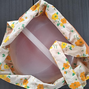 Può includere: Un contenitore rotondo in plastica traslucida con coperchio, inserito all'interno di una borsa di stoffa. La borsa presenta uno sfondo color crema con un motivo ripetuto di fiori giallo brillante e arancioni, insieme a foglie e steli verdi.