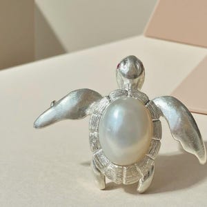 Può includere: Spilla a forma di tartaruga color argento con una grande perla lucente al centro. La spilla presenta ali a forma di pinna allargate e un design a guscio strutturato. Una piccola gemma rossa è incastonata sulla testa della tartaruga.