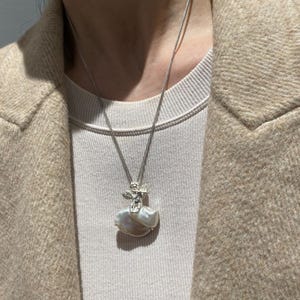以下が含まれることがあります： 小さな天使の姿が特徴のペンダントが付いたシルバーネックレス。大きな不規則な形の真珠の上にあります。真珠はクリーム色とタン色です。ネックレスはニュートラルな色調の背景に表示されています。