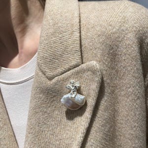 以下が含まれることがあります： 真珠を基盤とした銀色の天使のブローチが、ベージュのブレザーに留められています。ブローチは、広げた翼と流れるスカートを持つ天使の姿を特徴としています。ブレザーはクラシックなラペルとテクスチャード加工された生地です。